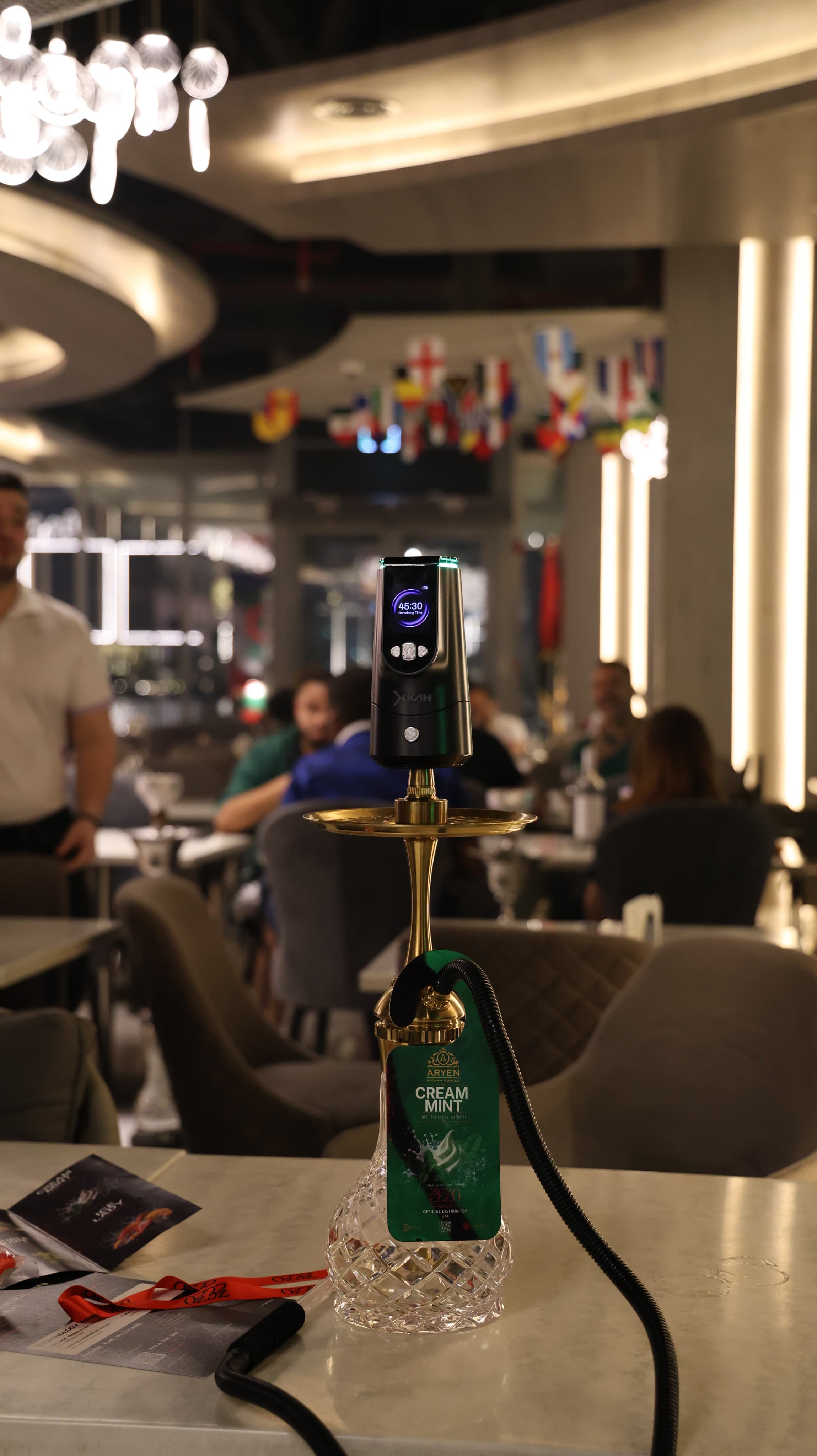 An XKAH heat manager on a gold-stemmed hookah at a table in a lit venue, guests seated behind